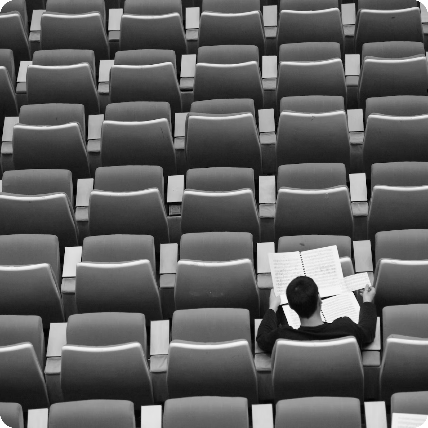 Imagen en blanco y negro de un hombre sentado en un auditorio vacío, luce un buzo y está leyendo gráficos en grandes hojas blancas